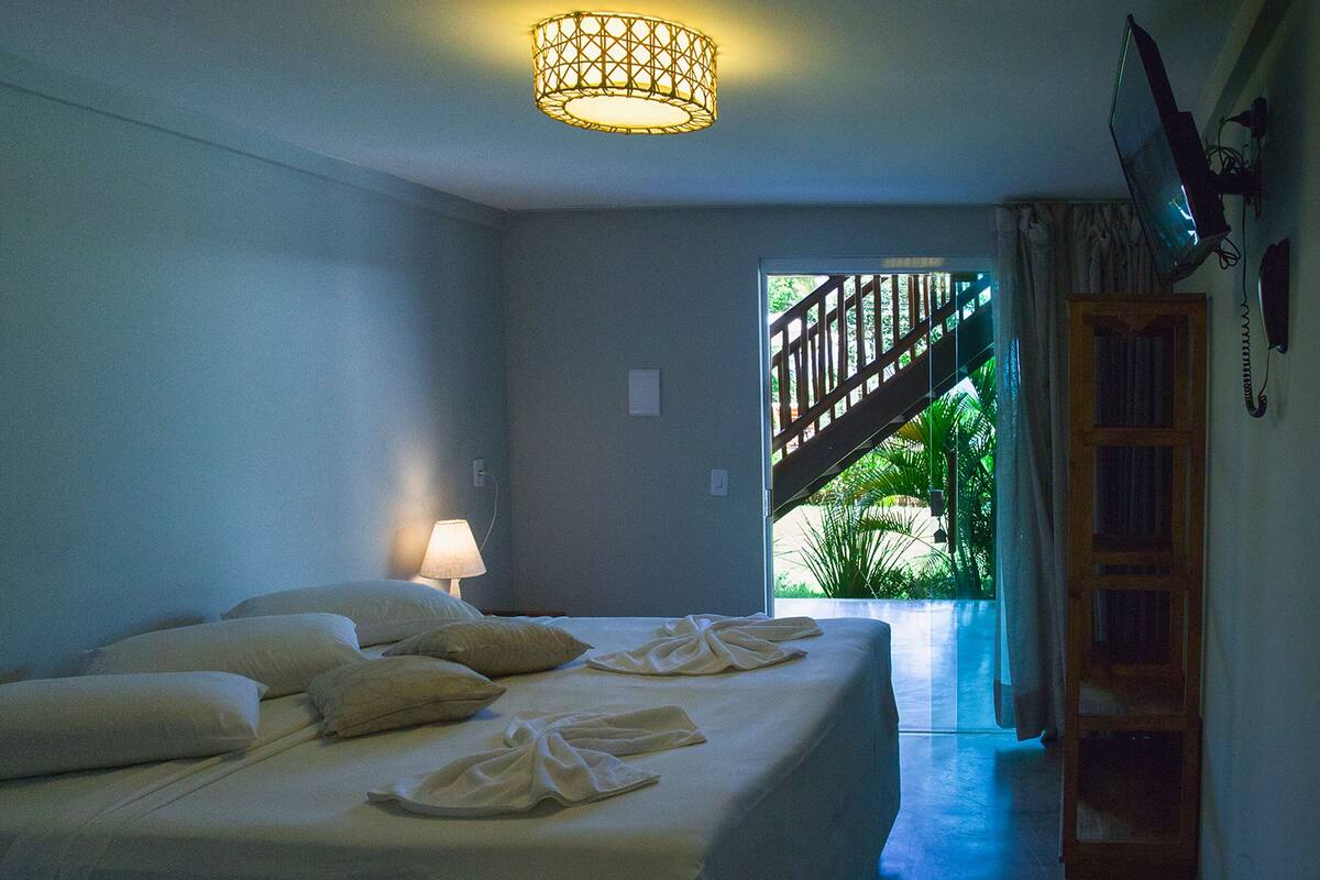 Quarto com cama, luminária de cabeceira, escada de madeira e vista para uma escadaria que leva a uma varanda.