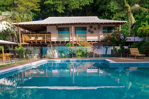 A pool with a wooden deck and lounge chairs, surrounded by lush greenery and a building with a tiled roof and wooden balcony railings. - Tropical Beach (Porto Seguro)