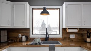 A modern kitchen with white cabinets, a black faucet, a wooden countertop, a kettle, and a window with a view of trees and a building.