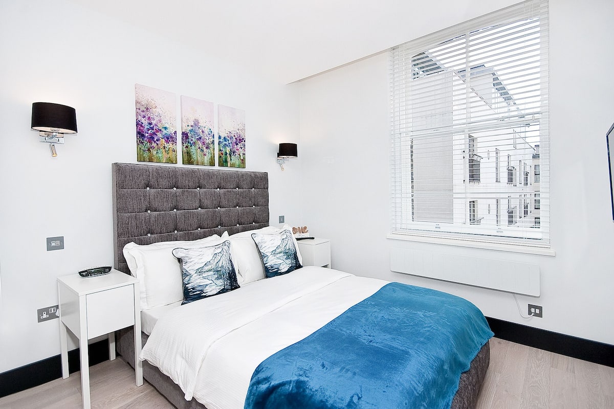 A modern bedroom with a grey tufted headboard, white bedding, and a blue throw blanket. There are two wall-mounted lamps, a nightstand, and a window with blinds.