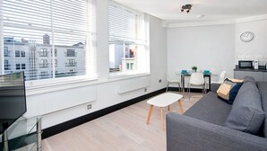 A modern living room with a grey sofa, a small white table, and a dining area with a white table and chairs. Large windows provide a view of the cityscape. - Heinze Building by City Living London (London)