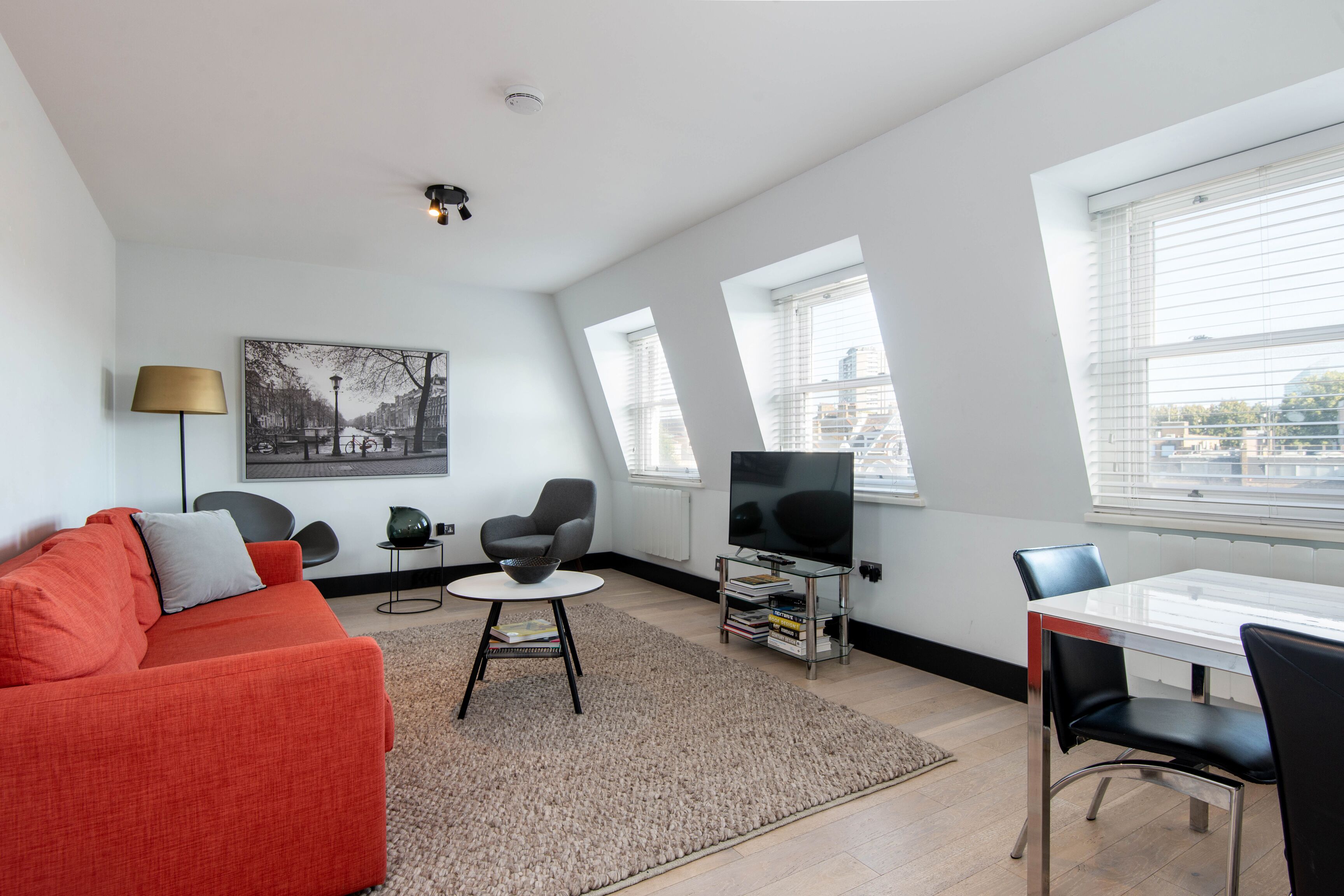 A modern living room with a red sofa, a black armchair, a small round table, a TV on a stand, and a dining table with chairs.