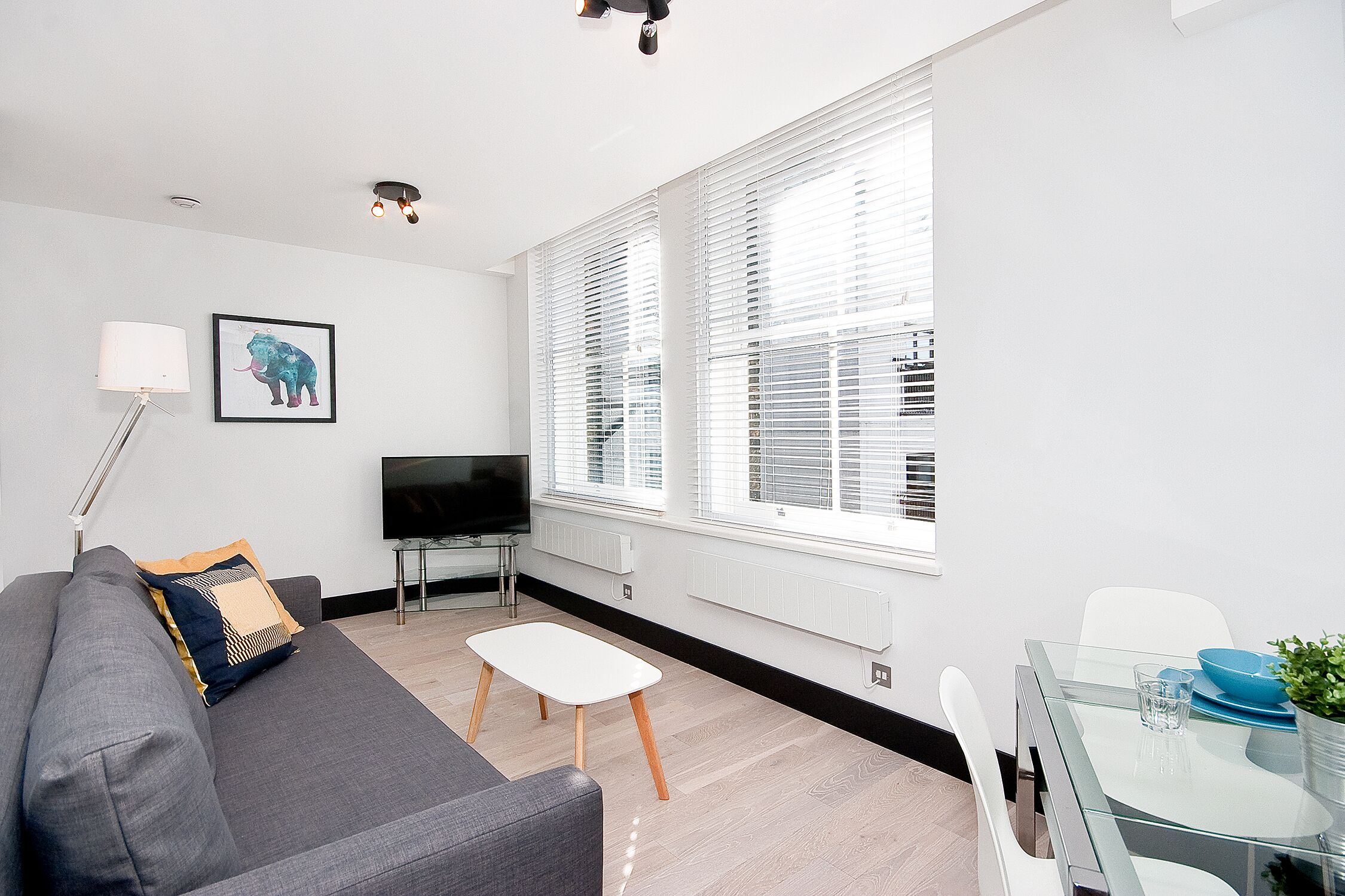 Ein modernes Wohnzimmer mit einem grauen Sofa, einem kleinen weißen Tisch und einem Glastisch mit Stühlen. Ein Flachbildfernseher ist an der Wand befestigt, und es gibt große Fenster mit Jalousien.