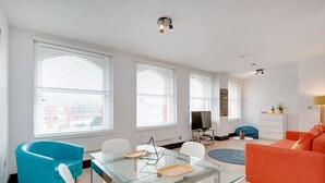 A modern living room with a glass dining table, a blue armchair, an orange sofa, and a television in the background. - Heinze Building by City Living London (London)