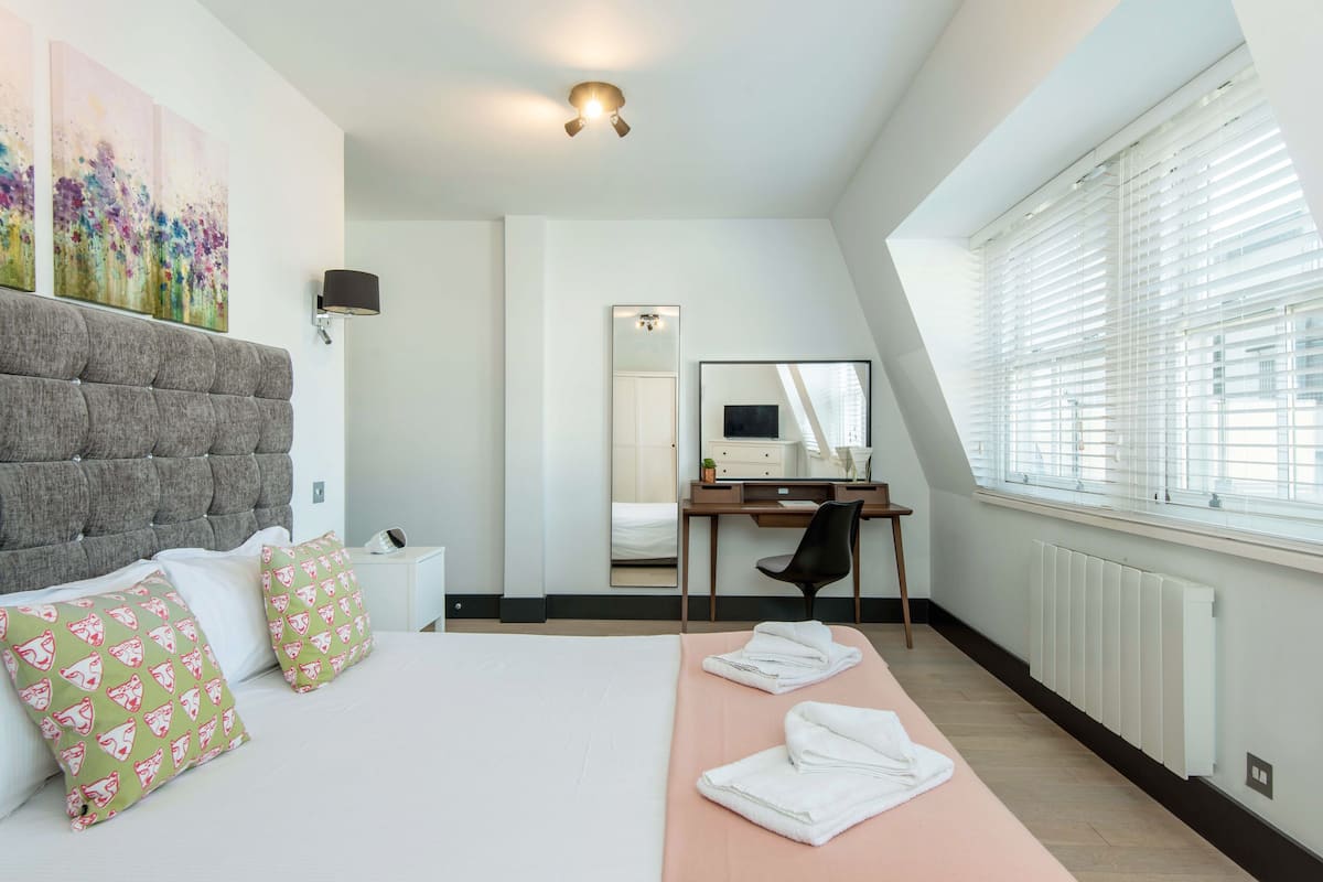 A modern hotel room with a large bed, a desk, and a window with blinds.