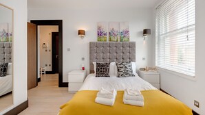 A modern hotel room with a large bed, a headboard with tufting, two bedside tables, a bedside lamp, a mirror, a window with blinds, and a hallway leading to another room. - Heinze Building by City Living London (London)