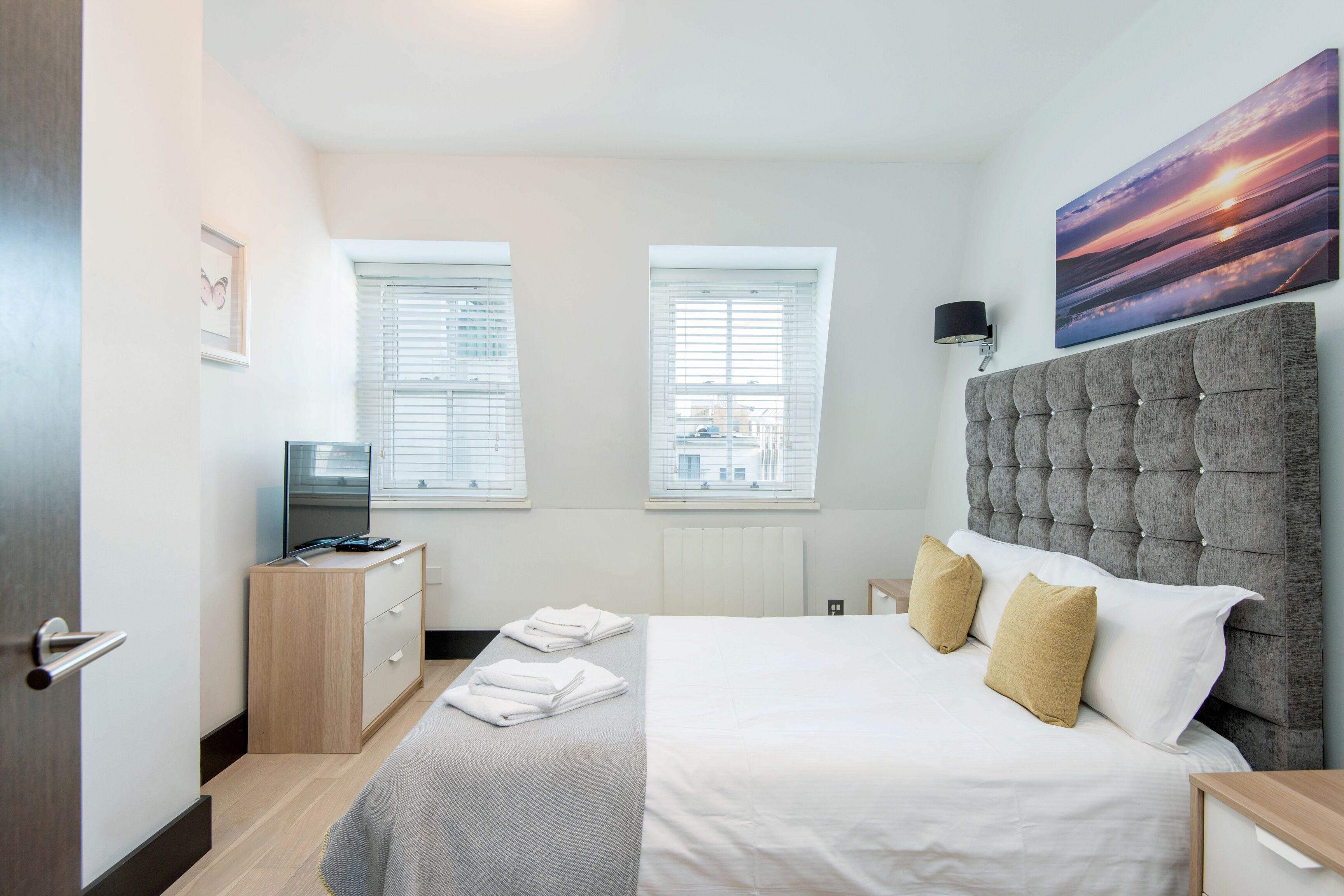 A modern hotel room with a large bed, a flat-screen TV, and two windows with blinds.