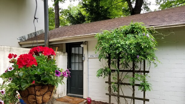 Une petite maison avec une porte noire, une treille en bois ornée de plantes grimpantes et un chemin de béton menant à la porte.
