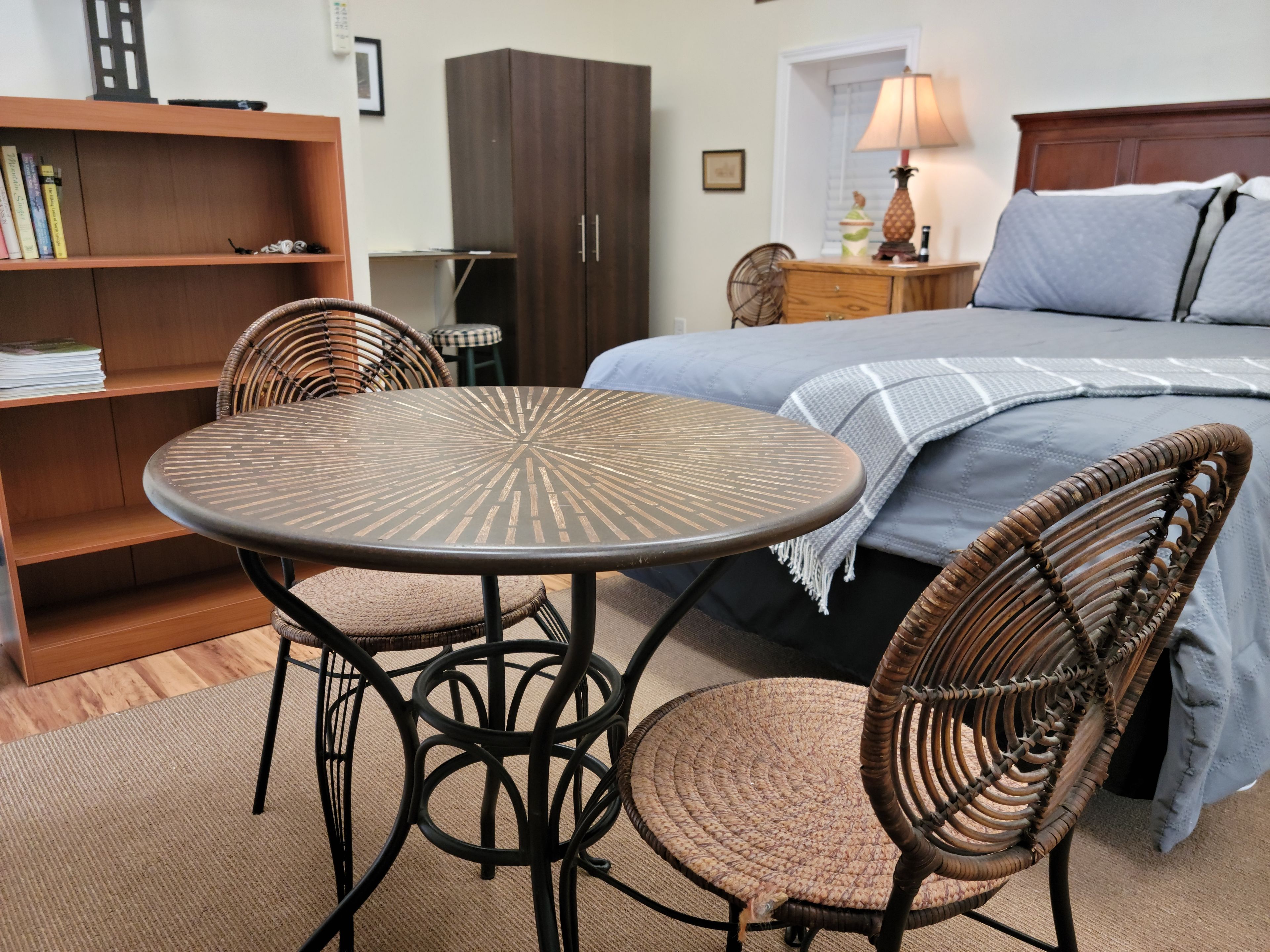 Une pièce comprenant un lit, une table ronde, deux chaises en osier, une bibliothèque et une armoire.