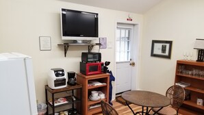 Une petite cuisine équipée comprenant un micro-ondes, un grille-pain et une machine à café, le tout placé sur une étagère. Un téléviseur est fixé au mur au-dessus, et un coin repas est aménagé avec des chaises en osier et une petite table ronde.