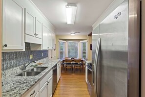 Une cuisine avec des meubles blancs, un plan de travail en granit, un réfrigérateur en acier inoxydable et un coin repas comprenant une table et des chaises en bois.