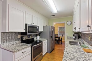 Una cocina con armarios blancos, electrodomésticos de acero inoxidable, microondas, horno, refrigerador, encimeras de granito, un reloj y una zona de comedor con mesa y sillas.