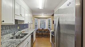 A kitchen with white cabinets, a granite countertop, a stainless steel refrigerator, and a dining area with a wooden table and chairs.