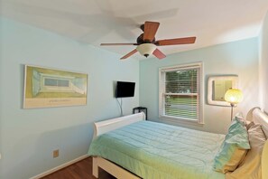 Un dormitorio con una cama, ventilador de techo, televisor montado en la pared, ventana con persianas y una lámpara de noche.