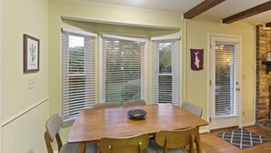 A dining area with a wooden table and chairs, white window blinds, a framed cactus artwork, and a stone fireplace.