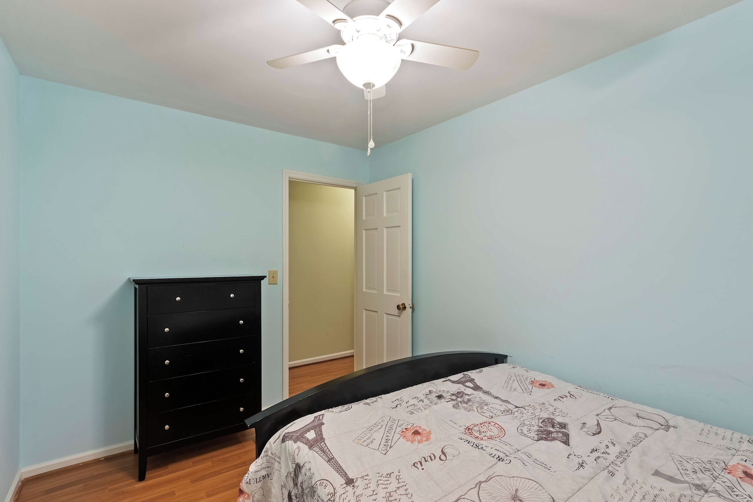 Une chambre à coucher comprenant un lit, une commode, un ventilateur de plafond et une porte donnant sur une autre pièce.