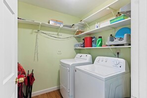 Un lavadero con dos lavadoras de carga frontal, estantes con productos de limpieza y un tendedero.