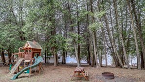 Une aire de jeux avec un toboggan et une structure d’escalade, une table de pique-nique et un foyer, le tout situé dans un espace boisé.