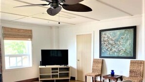 Un salon équipé d’un téléviseur à écran plat, de deux fauteuils en osier, d’une table basse en verre et d’un ventilateur de plafond.