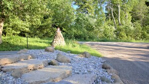 Un chemin de pierres mène à une construction en pierre surmontée d’un toit rouge, le tout étant entouré de verdure et d’arbres.