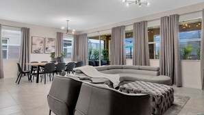 Una sala moderna con sofá seccional, comedor y ventanas amplias con cortinas.
