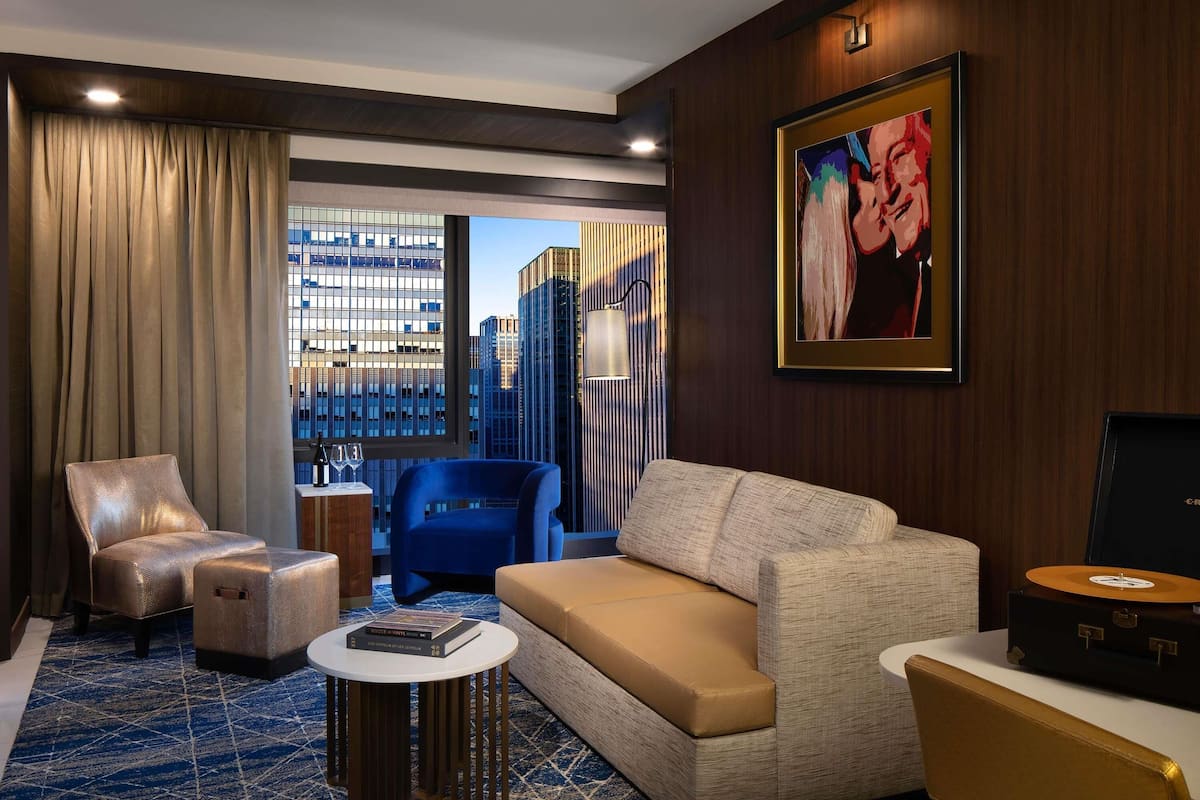 Una habitación de hotel moderna con un ventanal grande, un sillón azul, un sofá beige y una pequeña mesa redonda.