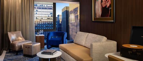 A modern hotel room with a large window, a blue armchair, a beige sofa, and a small round table.