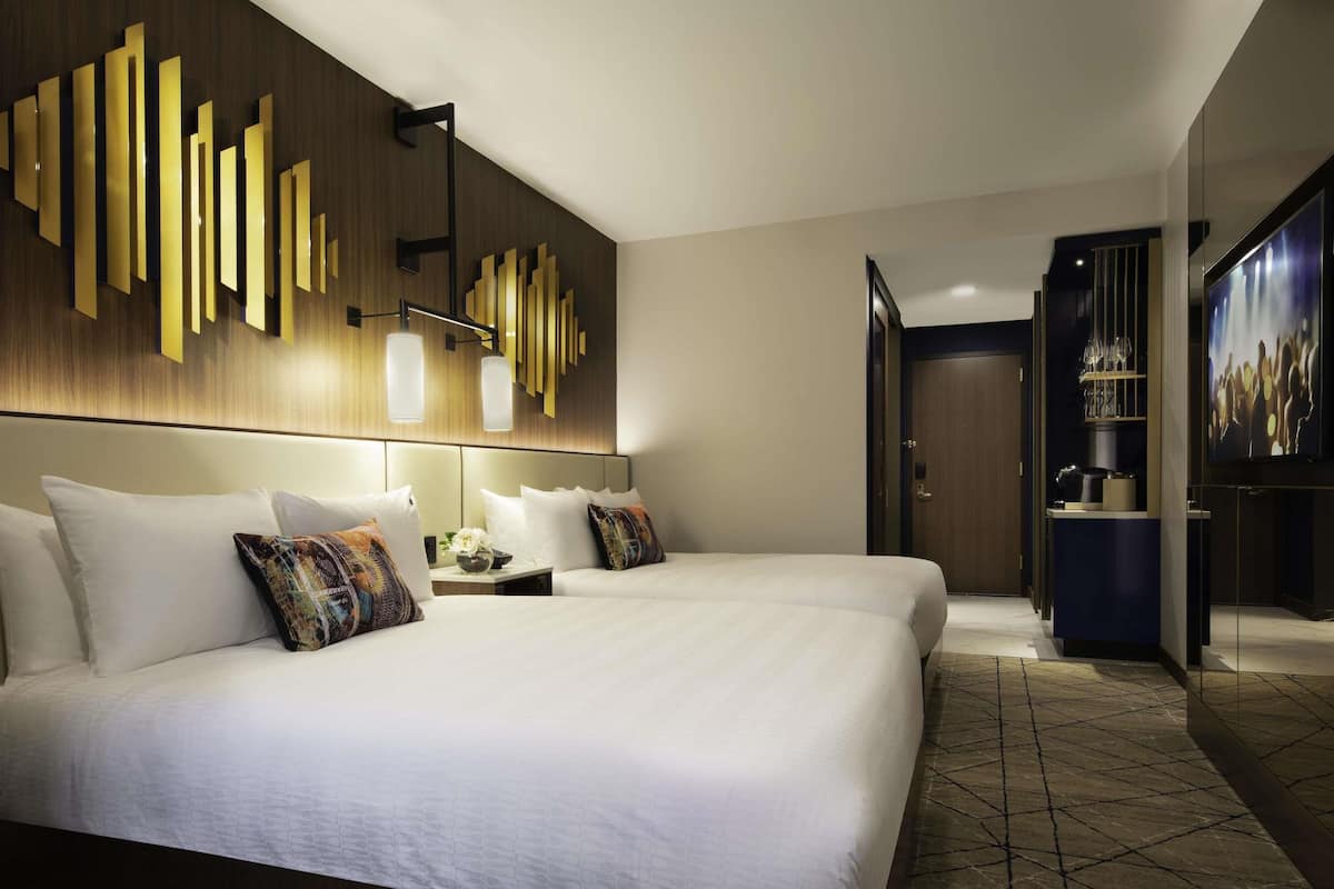 Una habitación de hotel con una cama grande, una mesita de noche con lámpara, un aplique de pared y una puerta que da al baño.