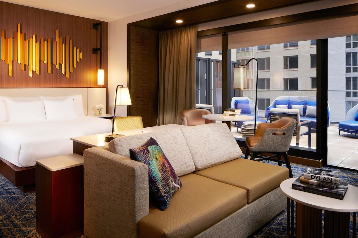 Habitación de hotel con cama, sofá, una mesita y vistas a la ciudad a través de amplias ventanas.