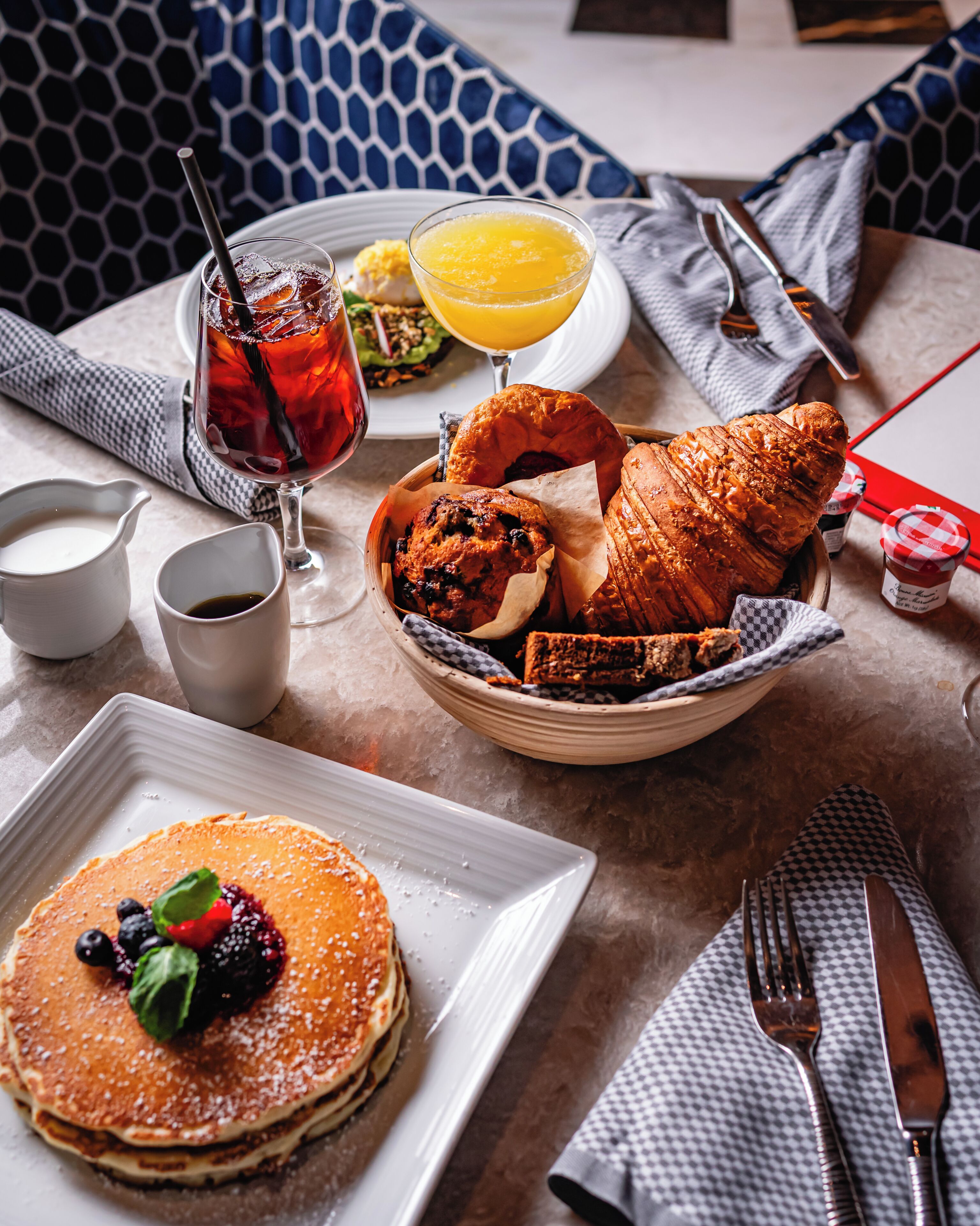 Mesa posta para o café da manhã com panquecas, croissants, muffins e um copo de suco de laranja.