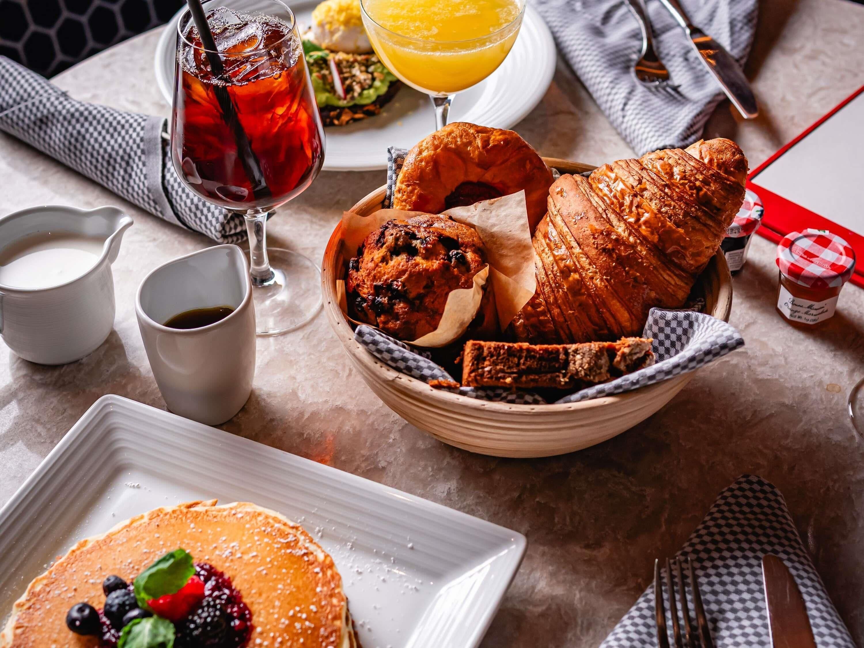 Mesa de café da manhã com um croissant, muffin e panquecas em um prato, um copo de suco de laranja e uma xícara de café.
