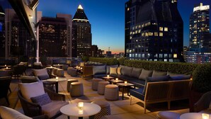 A rooftop lounge with outdoor seating, tables, and a city skyline view.