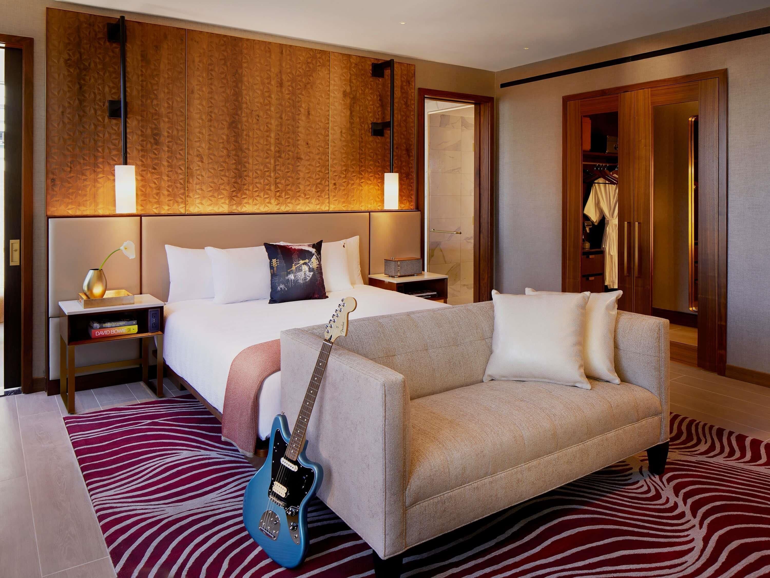 A hotel room with a bed, a sofa, a guitar, a nightstand, a lamp, and a wardrobe.