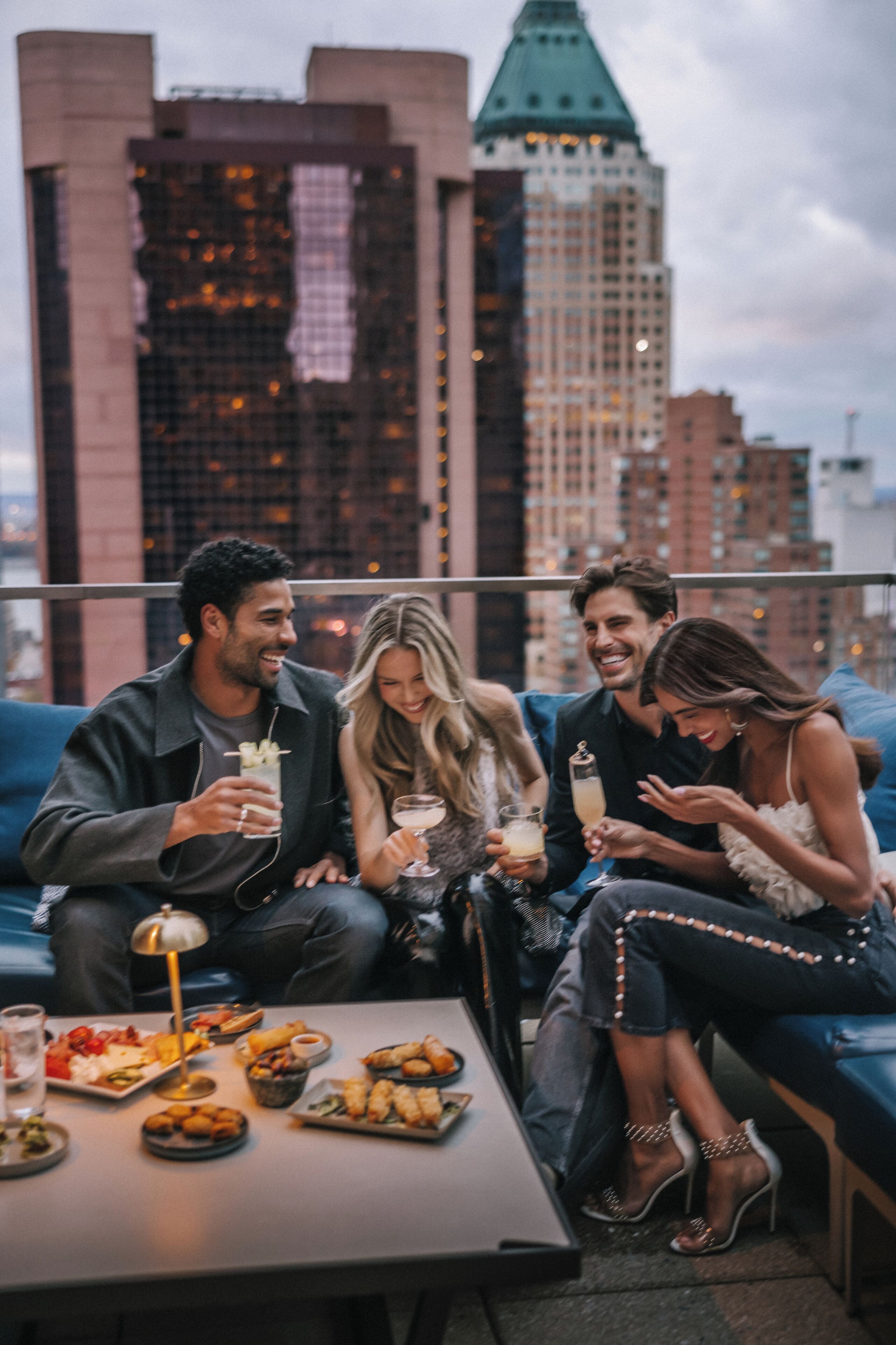 A group of people sitting on a blue couch with a table of food and drinks in front of them, with a cityscape in the background.