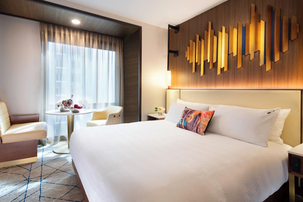 Habitación de hotel con una cama grande, un escritorio, una silla y vistas a la ciudad a través de amplias ventanas.