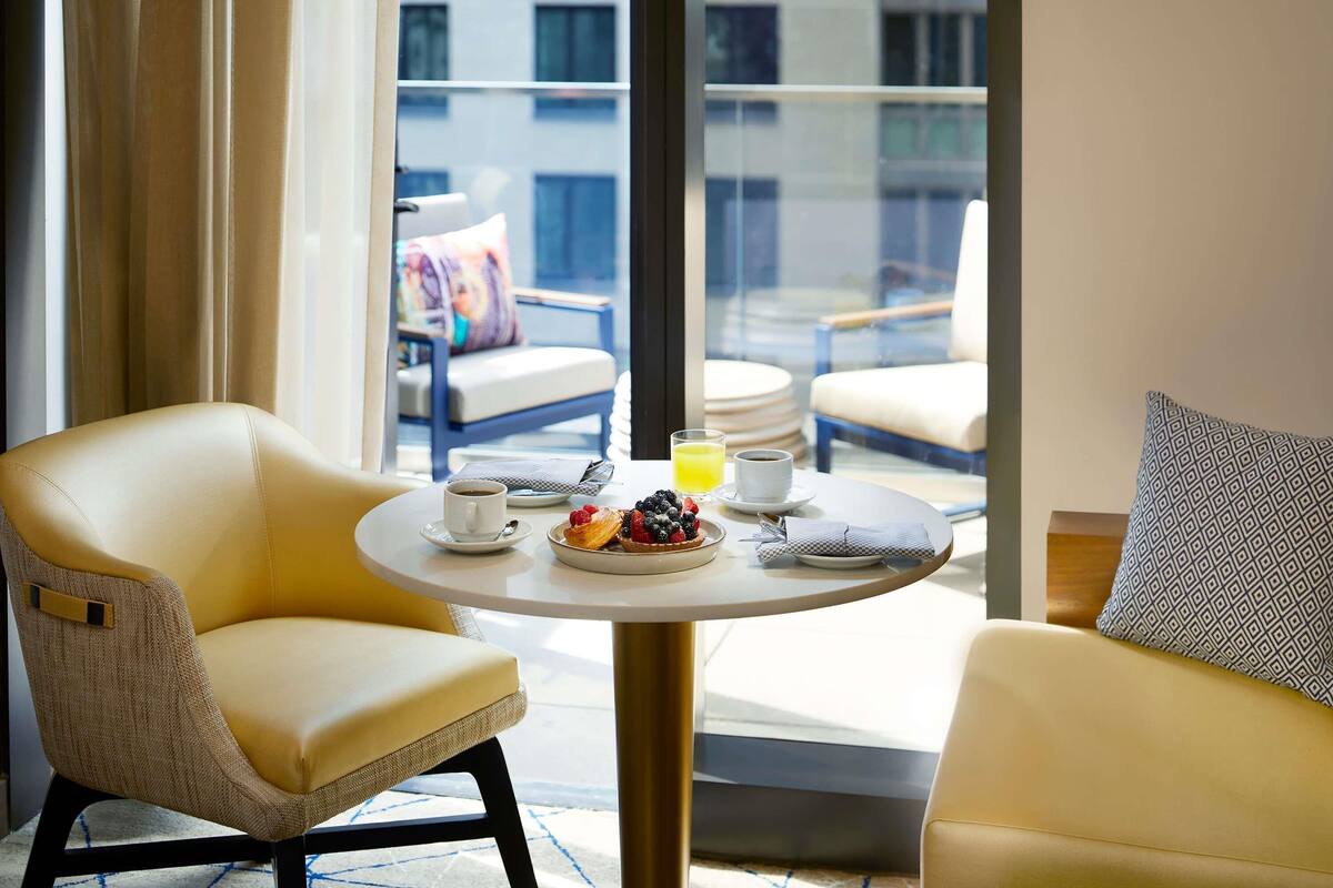 Habitación de hotel con balcón, dos sillas amarillas, una mesa redonda con desayuno y vista a edificios.