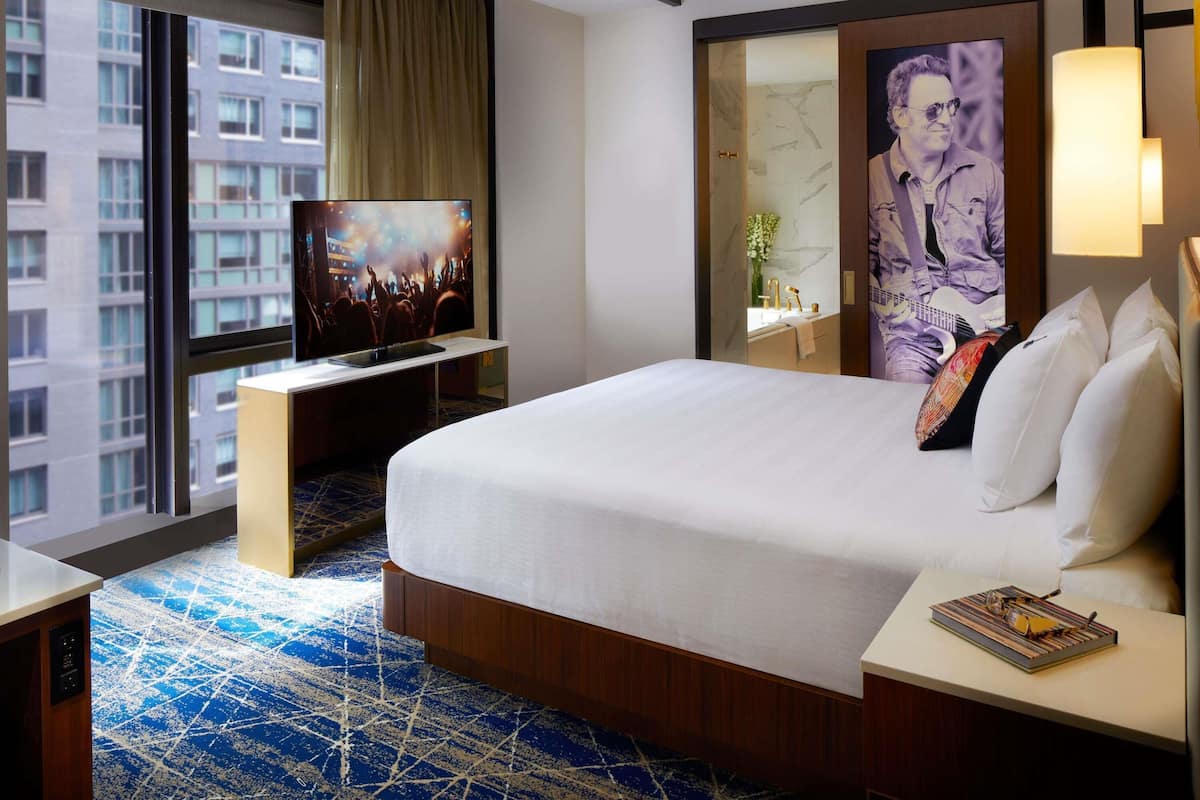 Habitación de hotel con una cama grande, un televisor sobre un mueble, un espejo de baño y vistas a la ciudad.