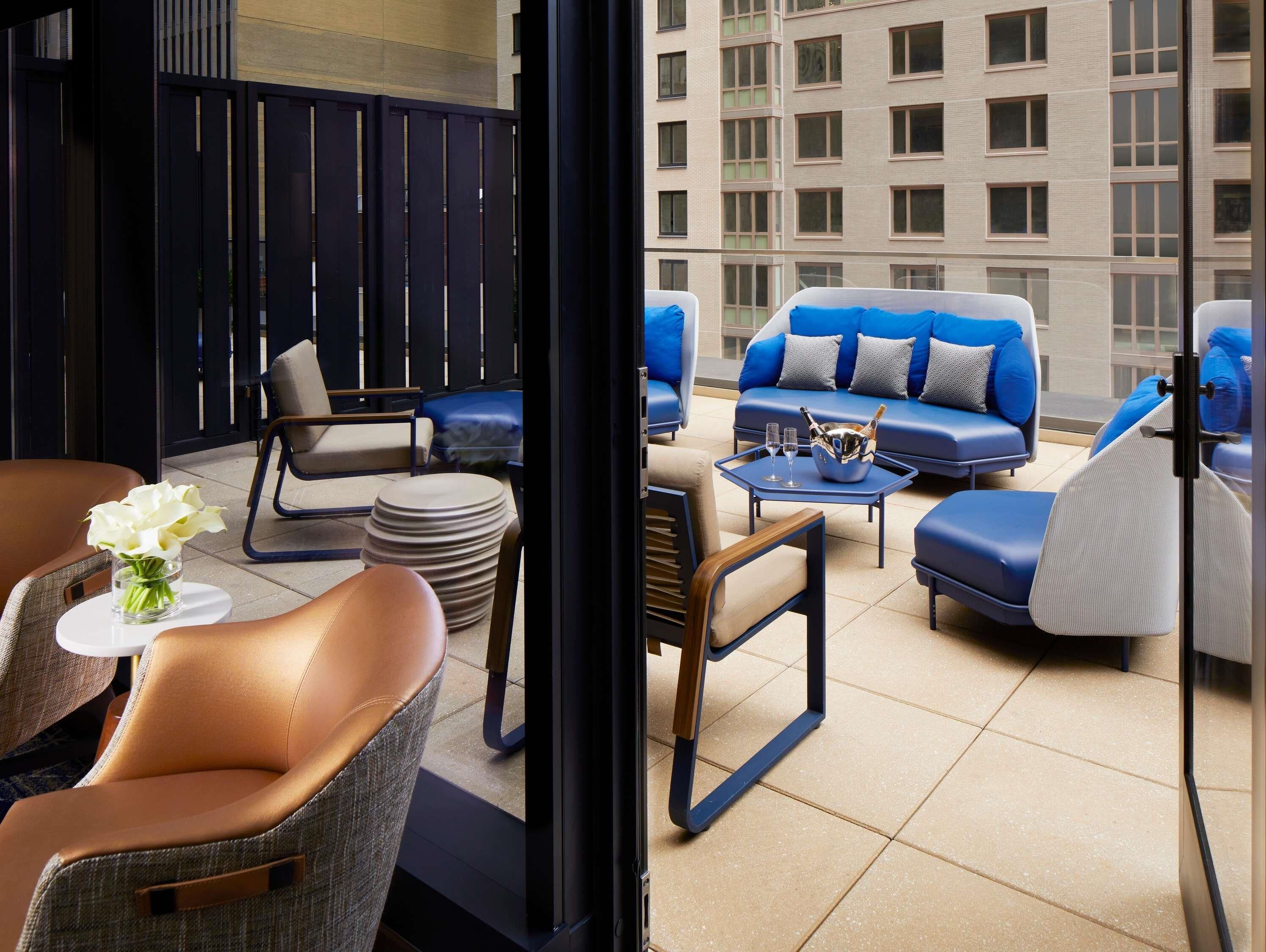 A modern lounge area with leather seating, a bar area with a bottle and glasses, and a view of a building with multiple windows.