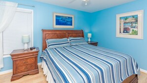Une chambre à coucher comprenant un lit, une table de chevet, une lampe, un ventilateur de plafond et deux tableaux encadrés accrochés au mur.