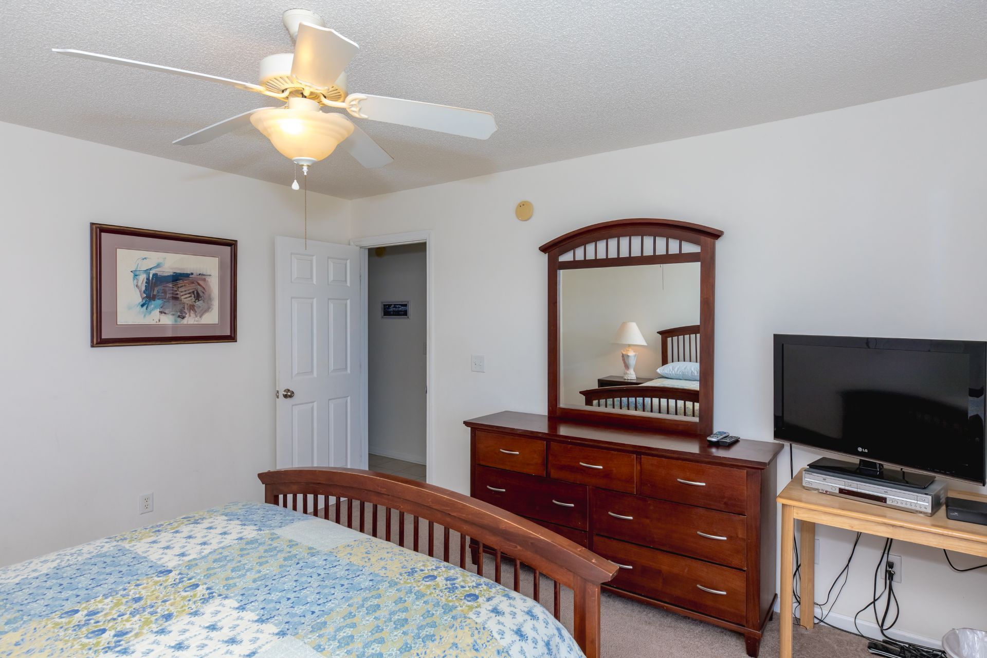 Une chambre à coucher comprenant un lit, une commode, un miroir, un téléviseur et un ventilateur de plafond.