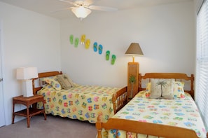 A bedroom with two single beds, a nightstand with a lamp, a ceiling fan, and a wall-mounted shelf with flip-flops.