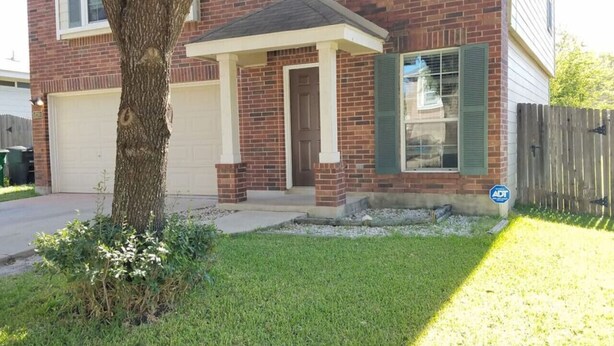 Una casa de ladrillo de dos plantas con garaje, un porche cubierto y un pequeño jardín delantero con un árbol y arbustos.