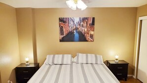 Habitación de hotel con cama, dos mesitas de noche, ventilador de techo y un cuadro de una escena callejera.