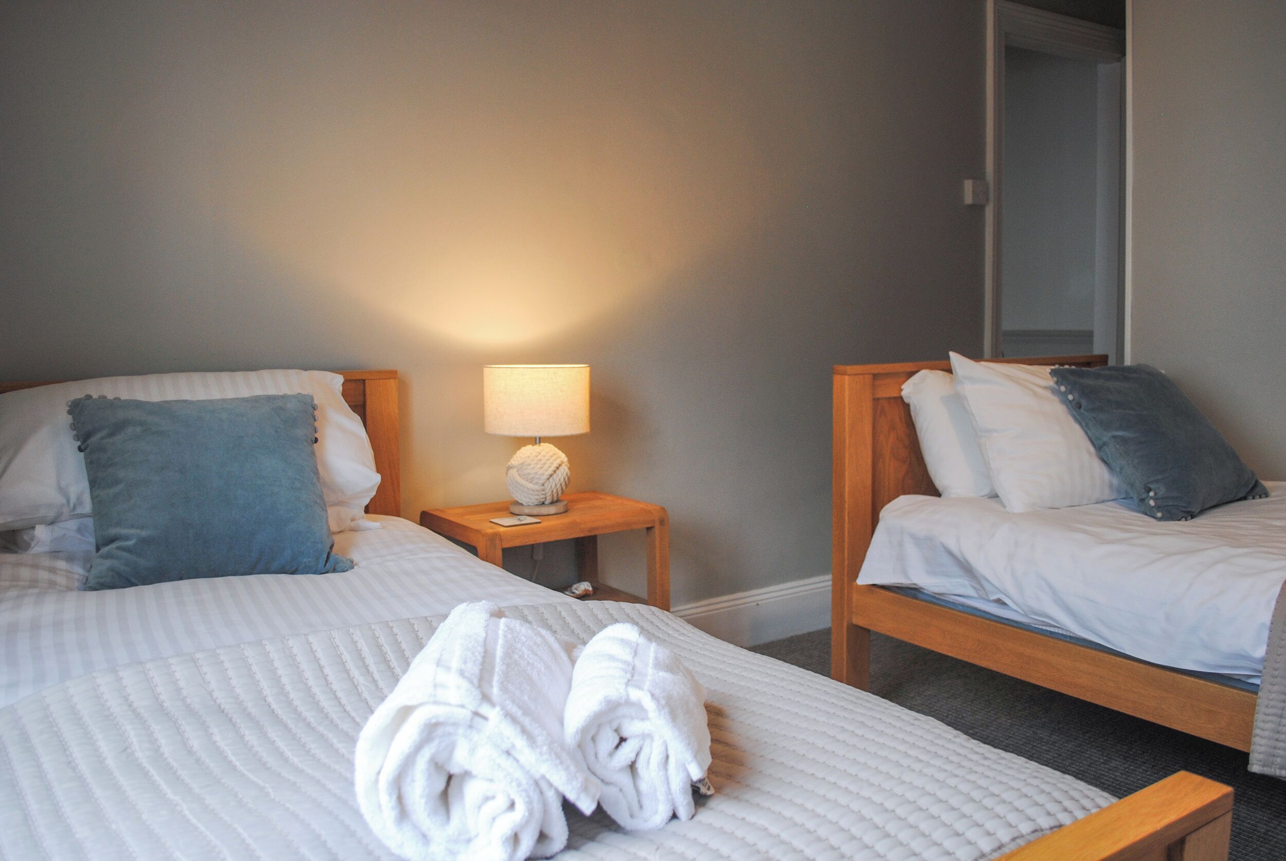 A hotel room with two beds, a nightstand with a lamp, and a rolled-up towel on the bed.