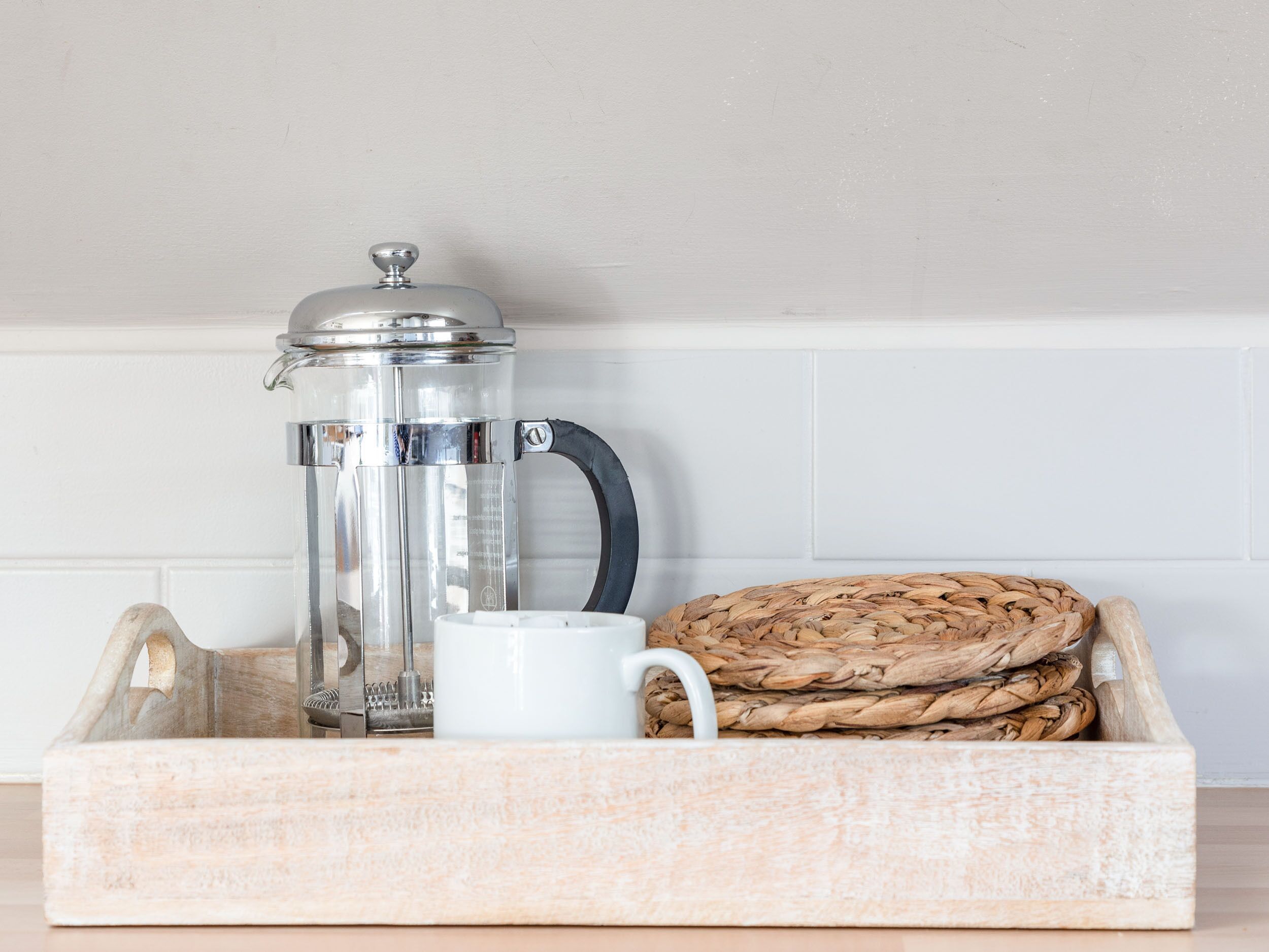 Une cafetière à piston en verre transparent, une tasse blanche et un panier tressé contenant divers objets disposés sur un plateau en bois.