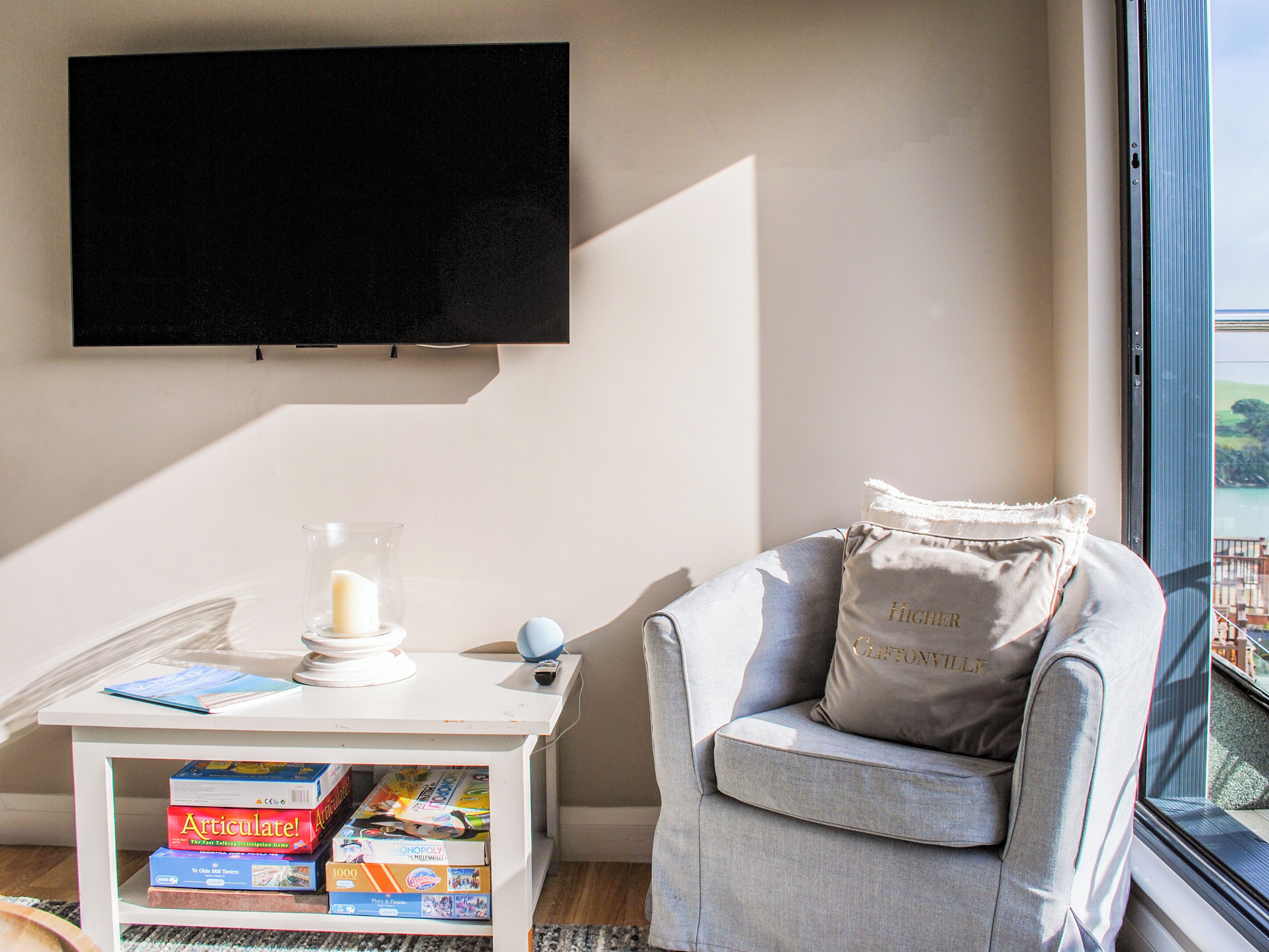 A living room with a flat-screen TV mounted on the wall, a small white side table with a candle and board games, a grey armchair with a cushion, and a view of a balcony through a window.