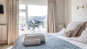 A bedroom with a large bed, a view of a village through the window, and a wall-mounted light fixture. - Higher Cliftonville (Dorking)