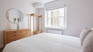 A bedroom with a large bed, a wooden dresser, a round mirror, a window with a view of a brick wall, and a small plant on the dresser. - 5VH Virginia House Cool Interiors in Bloomsbury 2 Double Beds sleeps 4 (London)