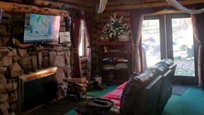 Ein Interieur einer Blockhütte mit Kamin aus Stein, einem an der Wand befestigten Fernseher, einem Ledersofa und einem großen Fenster mit Vorhängen.