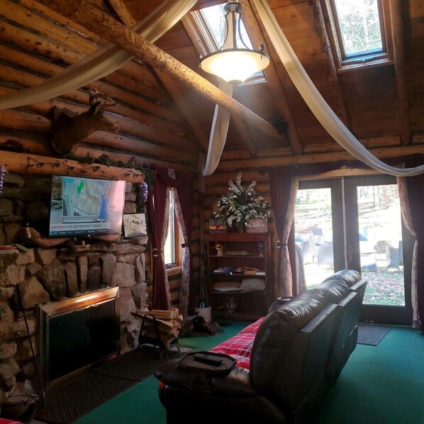 A log cabin interior with a stone fireplace, a television mounted on the wall, a leather couch, and a large window with curtains.